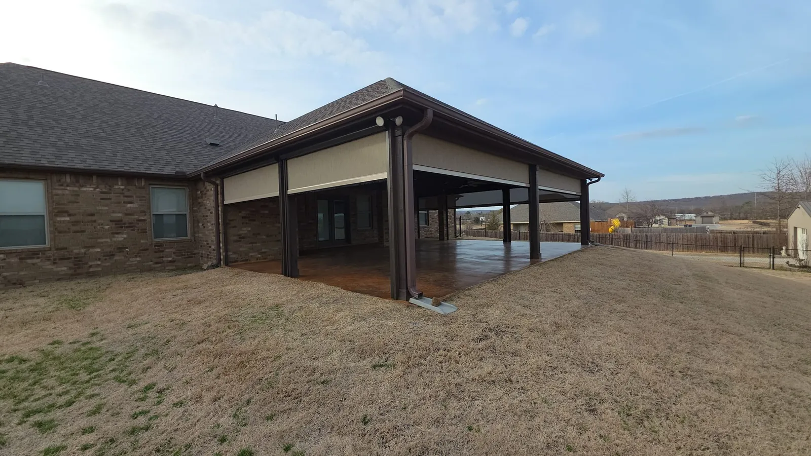 Covered back patio in Bentonville, Arkansas with motorized retractable screens partially lowered, brick home with stained concrete patio, screen installation by NWA Security Shutters