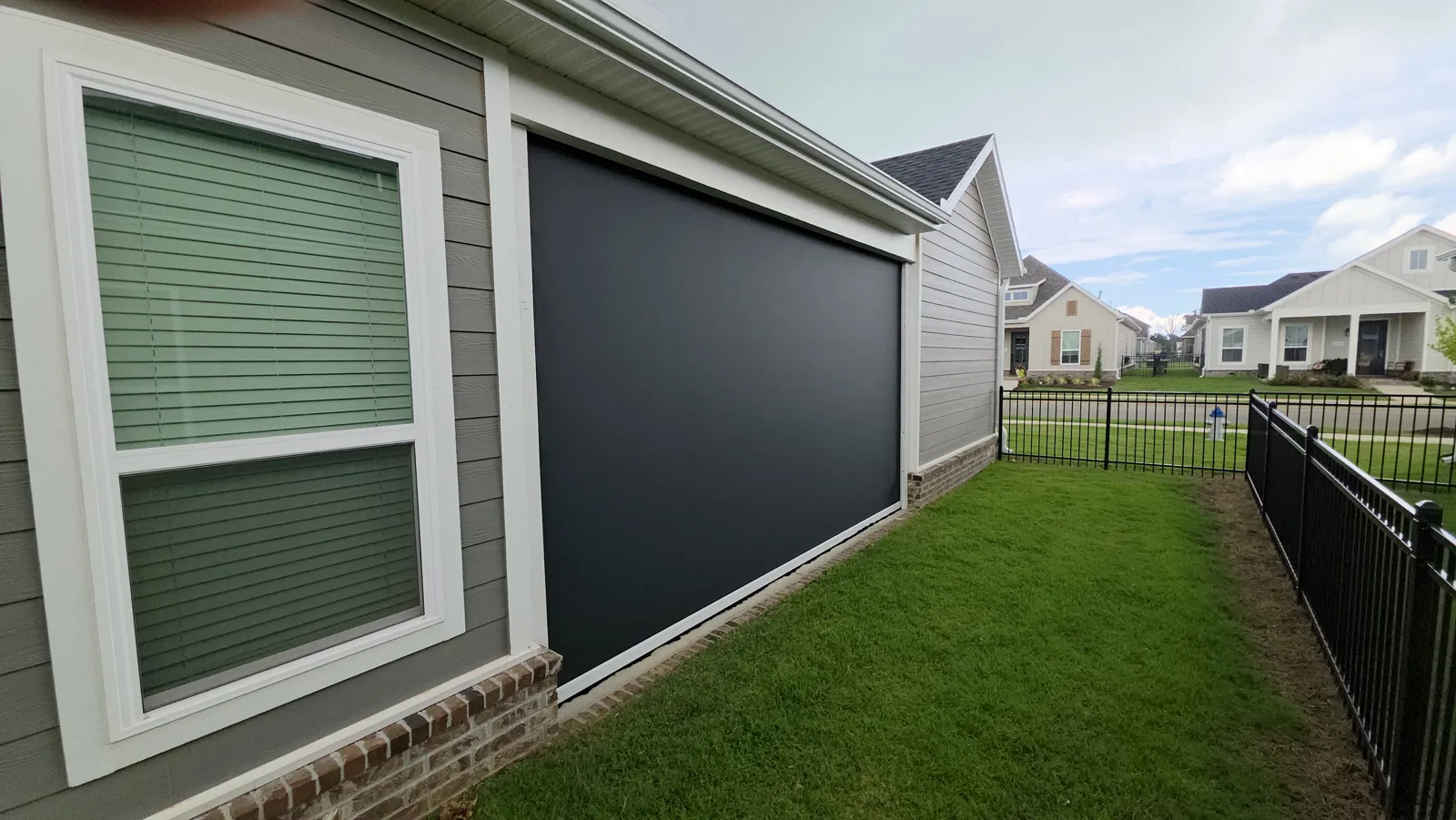 A dark screen closed beside a window on a gray-green sided house with a brick base, with a black iron fence and lawn, retractable screens for homes in Ridgecrest Meadows, Rogers, Arkansas