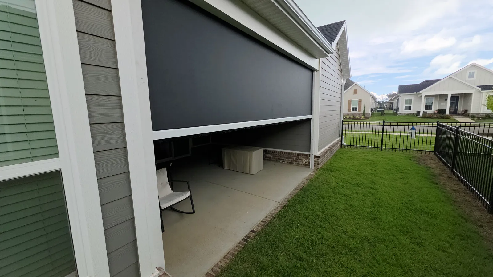 A dark screen partly lowered on a covered patio of a gray-green sided house, with a black iron fence and lawn, retractable screens for homes in The Enclave, Springdale, Arkansas