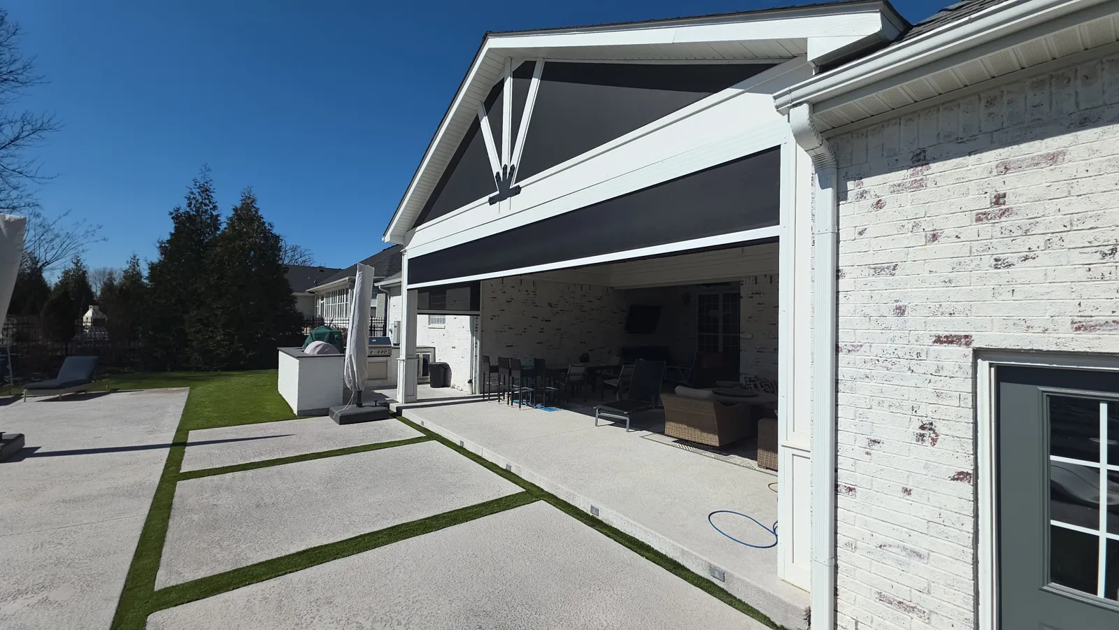 A dark screen closed under a gabled patio on a white painted brick house, with a stamped concrete patio and trees, retractable screens for homes in Ivey's, Rogers, Arkansas