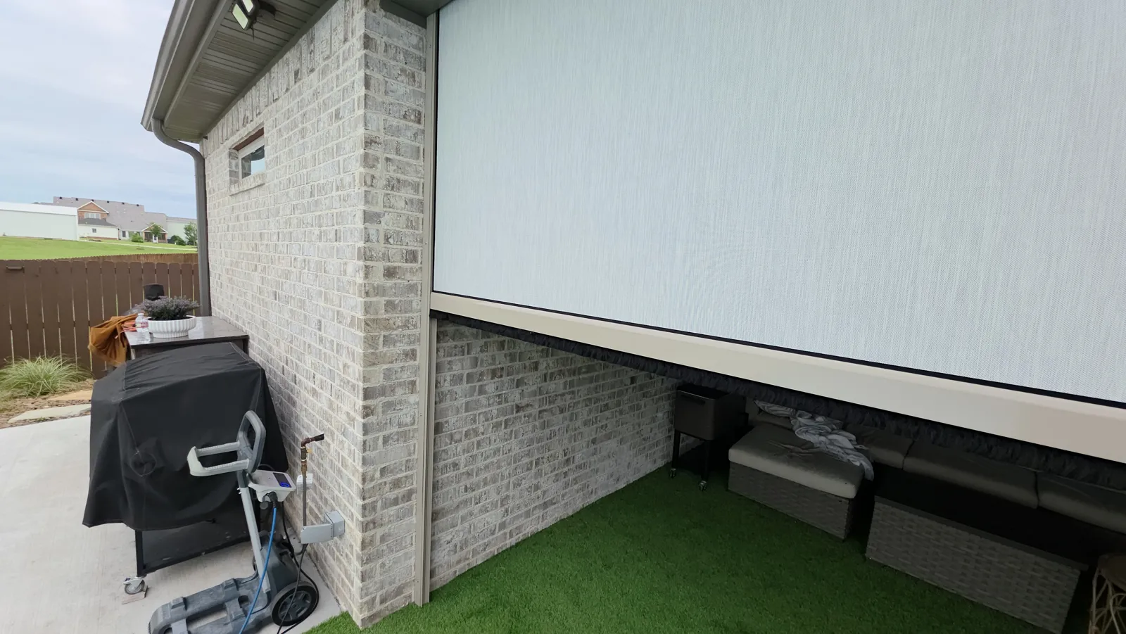 A light gray screen closed on the side of a brick house over a covered patio, with a grill and artificial turf, retractable screens for homes in Wilkins Estates, Lowell, Arkansas