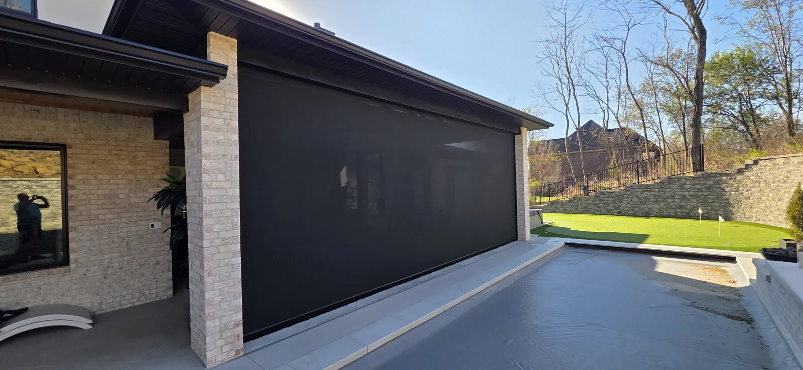 A dark screen closed beside a brick column on a house with a tan brick wall, looking out to a wet patio and a lawn, retractable screens for homes in Huffman, Bentonville, Arkansas