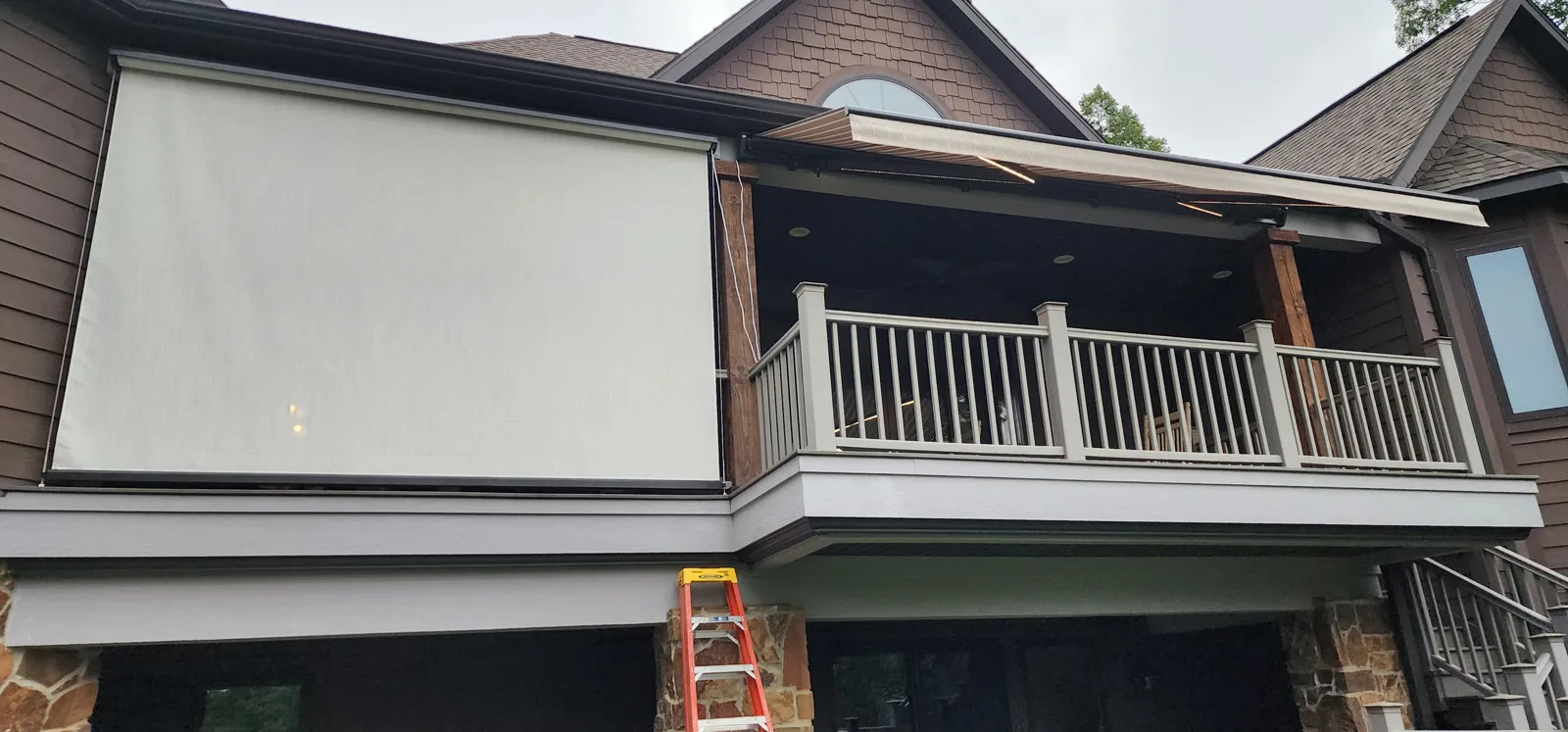 A light cream screen closed over an upper deck with a white railing on a brown sided house, with a stone base and a ladder, retractable screens for homes in Bliss Meadows, Centerton, Arkansas