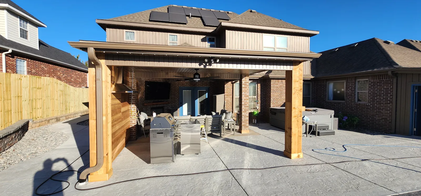 A cedar-post patio cover with a ceiling fan and an outdoor kitchen behind a brick two-story house, with solar panels on the roof, retractable screens for homes in Garner, Lowell, Arkansas